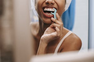 Young African American woman practicing oral hygiene by flossing teeth in front of a bathroom mirror. | Dentists Hate This: 8 Flossing Secrets They’ll Never Tell You (Number 5 Is Gross)