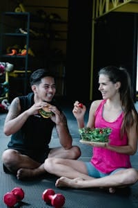 A couple laughs while sharing a salad and burger sitting on a gym floor. | Deflate Your Belly Fast: 10 Science-Backed Superfoods That Shrink Bloat & Get You Flat!