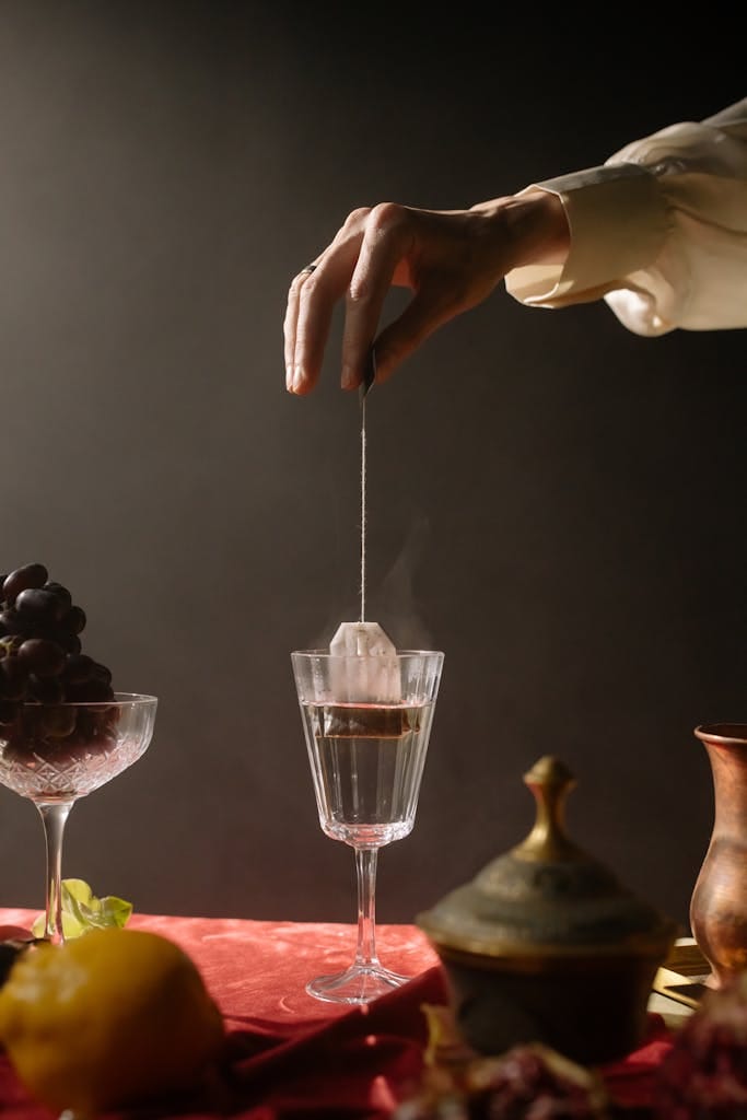 A serene setting with a hand placing a teabag in a steaming glass, surrounded by fruits and vintage glassware. | Constipated? These 5 Drinks Are Your Instant Bathroom Pass (Seriously!)