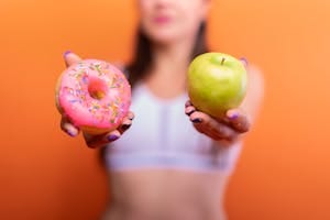 A woman holds a green apple and a pink donut with sprinkles against an orange background, emphasizing choice. | Dietitian's Urgent Warning: 7 Inflammatory Foods Secretly Fueling Your Visceral Fat (Cut These NOW!)