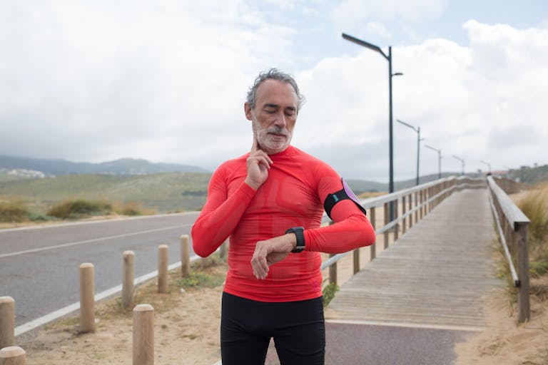 Elderly man in red activewear checking pulse on a coastal jogging path. | longevity exercise