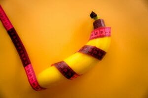 Conceptual image of a banana wrapped with a pink measuring tape on a bright yellow background. | placeholder used to illustrate Ozempic Penis