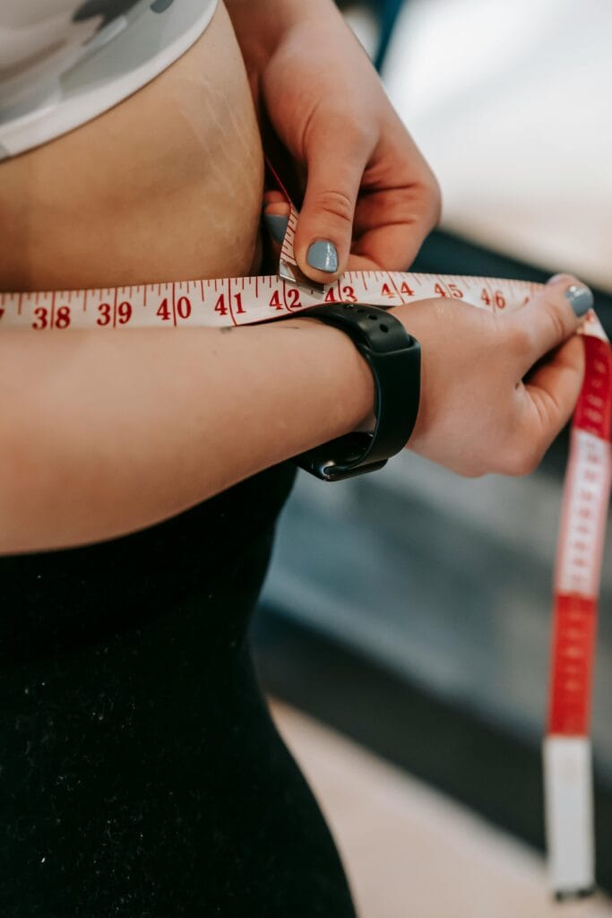 Crop anonymous female with measuring tape around big belly on blurred background of gym | Wake Up Slimmer: 4 Morning Habits After 50 That Attack Belly Fat, No Workouts