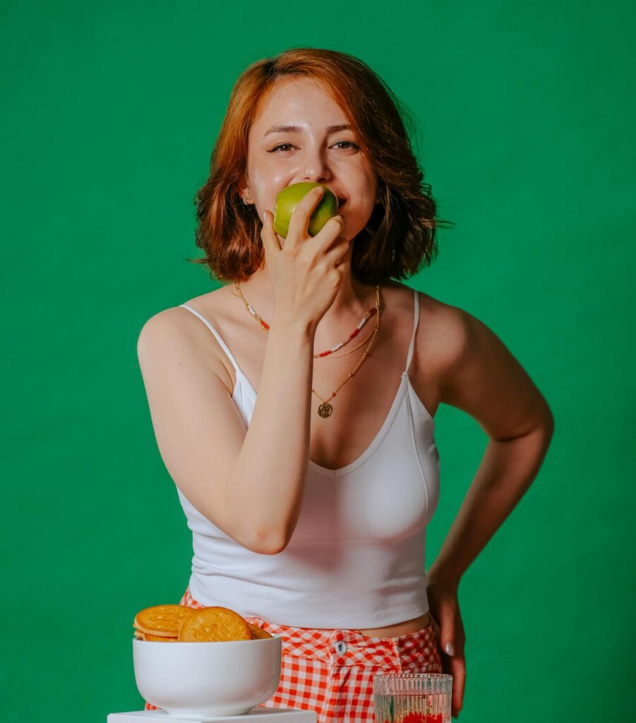 Smiling woman eating an apple, surrounded by vintage kitchen decor, creating a cheerful retro atmosphere. | Ozempic-Level Results? Unlock Your Body's OWN Fat-Burning Hormones By Eating Smarter (Science-Backed!)