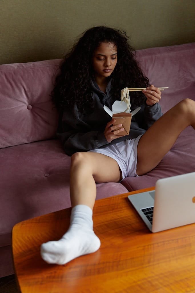 Woman sitting casually on a sofa, eating takeout noodles with a laptop nearby. | The 7 Foods Dietitians Say Are Secretly Ruining Your Thyroid (And What to Eat Instead)