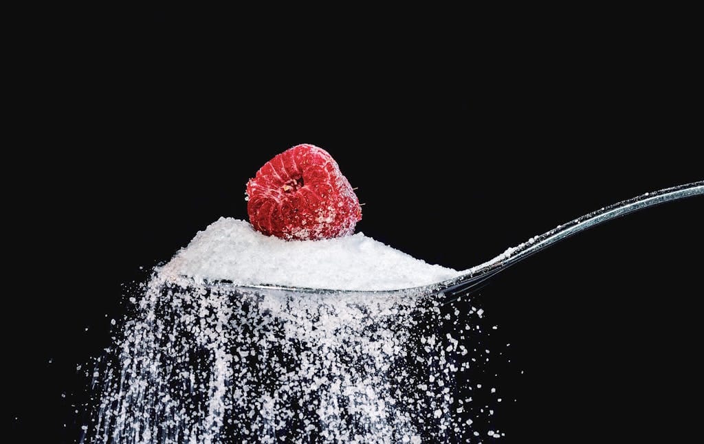 A vibrant raspberry resting on a sugar-coated spoon against a black backdrop. | Sweet Victory: 7 Natural Sweeteners That Taste Incredible & Keep Your Blood Sugar Flat