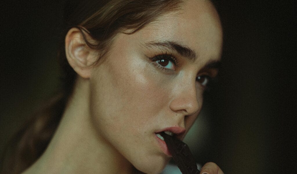 An artistic close-up portrait of a woman biting a chocolate bar, capturing a thoughtful expression. | Chocolate, Cheese & Coffee? Doctors Launch NEW Gut Health Diet You Won't Believe!