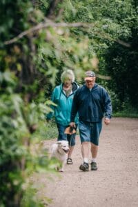 An elderly couple strolling with their pet dog in a scenic green park on a pleasant day.