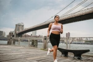 A woman jogging near the iconic Brooklyn Bridge with a cityscape background. | The Desk Job Antidote: Experts Reveal How Much Exercise Truly Erases A Full Day of Sitting