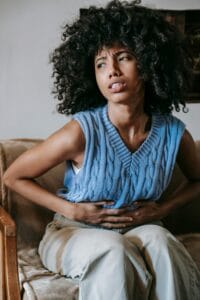 Young African American female sitting on couch at home while having acute pain in stomach | GI Doctor's Warning: This Common Food Secretly Triggers Uncontrolled Bowel Leaks