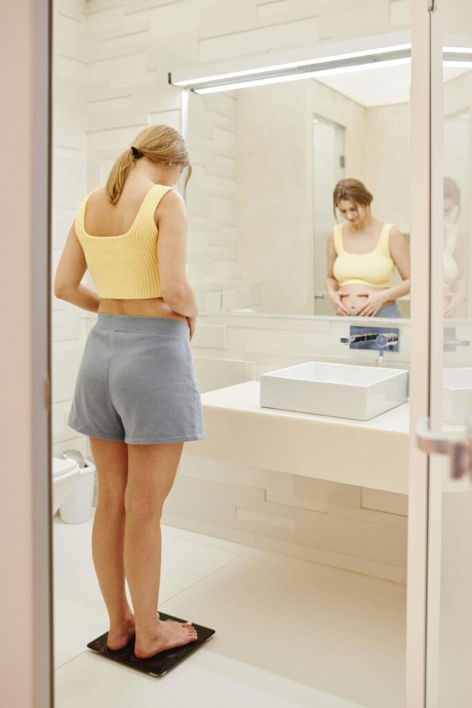 Pregnant woman standing on a scale in a well-lit modern bathroom, a moment of anticipation. | Stop Scale Stress: You're Probably Weighing Yourself All Wrong. Here's When to Step On