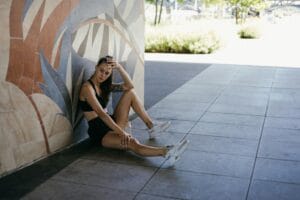 A young woman in sportswear rests by a vibrant mural outdoors, looking contemplative. | You're Dehydrated & Don't Know It: This Secret Is Aging Your Body FAST! (The Simple Fix)