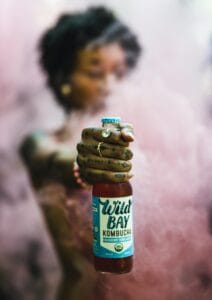 Close-up of a tattooed hand holding a Wild Bay Kombucha bottle with a misty background. | Drink Kombucha, Fight Fat: New Study Reveals Gut Microbes Melt Pounds (No Diet Needed!)