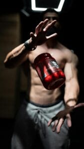 A shirtless man skillfully tosses a supplement jar in a dramatic indoor setting, showcasing fitness and agility. | Summer Workout Revolution: The TOP Supplements to Maximize Your Gains (Beat the Heat!)