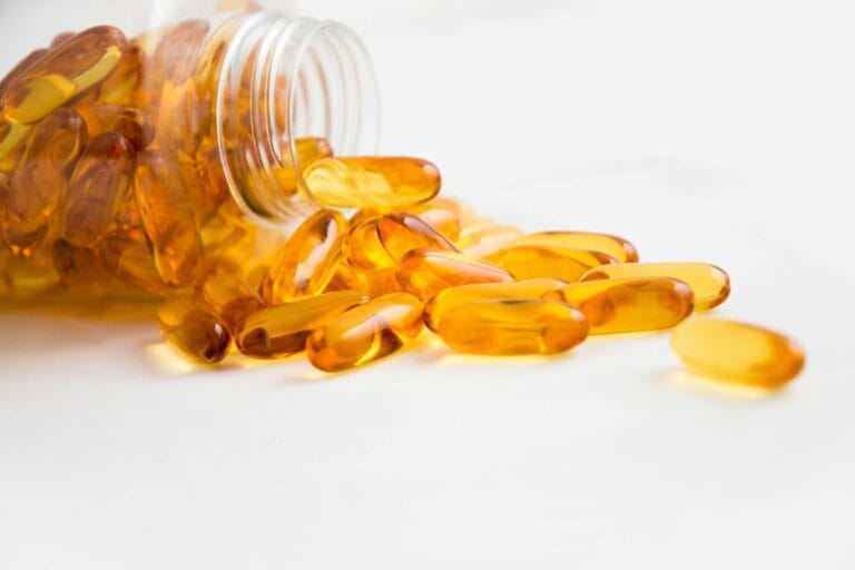 Close-up of fish oil capsules spilling from a bottle on white background. | Omega-3 Side Effects You’re Not Hearing About—Doctors Say Watch Out for These