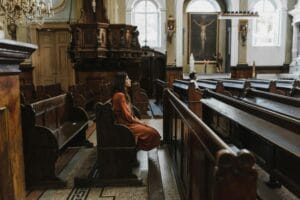 A woman sits quietly in a chapel, surrounded by wooden pews and religious decor.