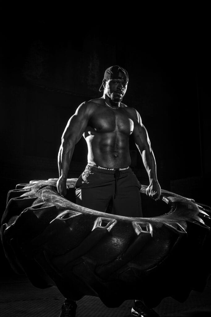 A muscular bodybuilder lifting a heavy tire in a dramatic black and white capture. | Want Bigger Gains in Less Time? This Workout Hack Cuts Your Gym Time by 40%
