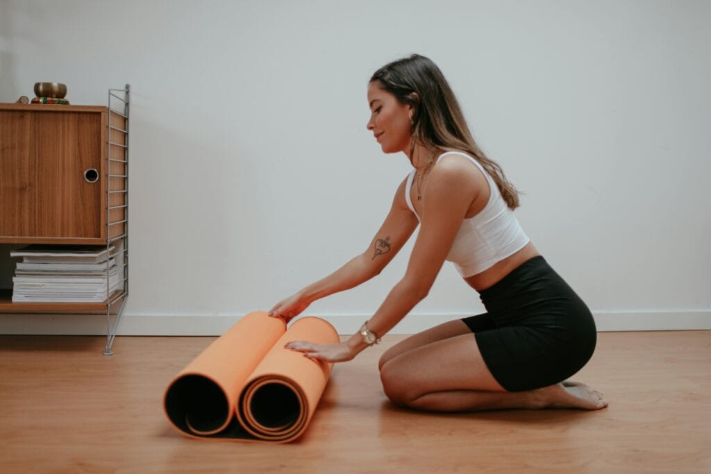 A woman rolls up an orange yoga mat on a wooden floor, ready for a yoga session.