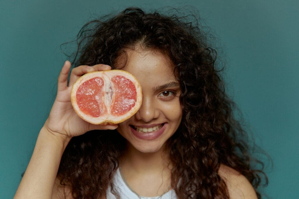 Young woman with curly hair smiling and holding a grapefruit half over her eye. | This Overlooked Fruit Could Be the Key to Balanced Blood Sugar—Longevity Experts Are Talking