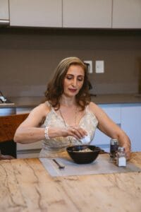 Elderly woman seasoning a dish in a kitchen setting, adding flavor to her meal. | Defy Your Aging Gut: An Expert's 5-Step Plan to Revitalize Your Microbiome & Feel Younger