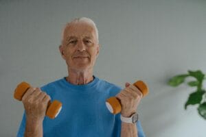 Elderly man in blue shirt lifting dumbbells and smiling indoors, promoting active lifestyle. | 10 Powerful Exercises That Fight Frailty and Keep You Moving Strong After 50
