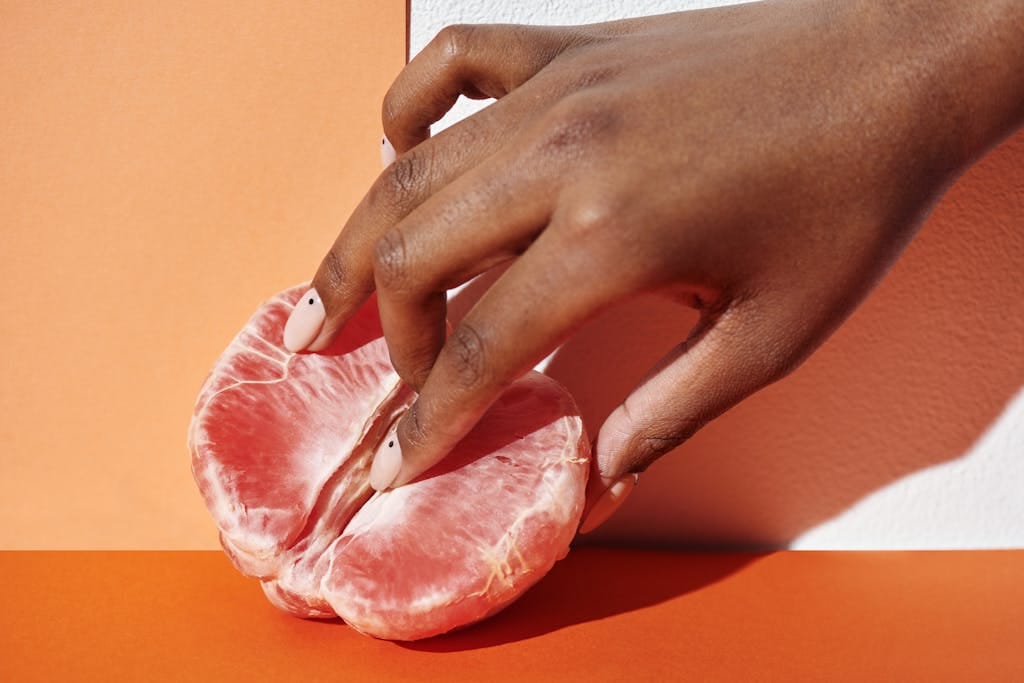 Close-up of a hand gently touching a grapefruit against an orange background, showcasing freshness and elegance. | The ‘Ozempic Vulva’ Trend: What Rapid Weight Loss Is Doing to Women’s Genitals—and Why Some Are Turning to Surgery