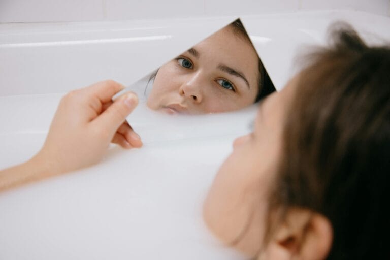 A woman lies in a milky bath, holding a mirror reflecting her serene expression.