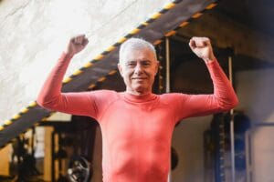 Elderly man in gym raising arms in celebration wearing a red top.