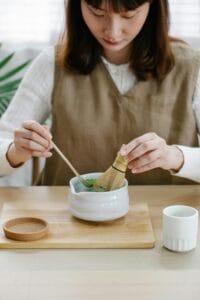 Asian woman making matcha tea with whisk, highlighting traditional preparation.