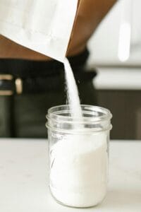 Close-up of person pouring sugar into a glass jar on a kitchen counter, showing organization. | Forget the Injections—Doctors Are Buzzing About This Sugar Alternative That Could Mimic Ozempic for Fat Loss
