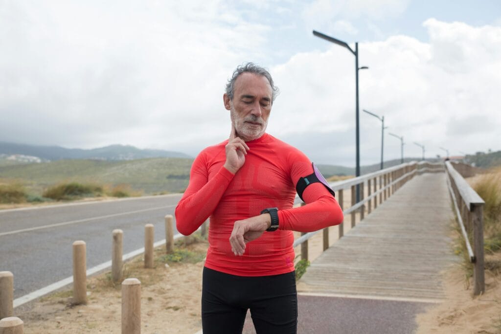 Elderly man in red activewear checking pulse on a coastal jogging path.