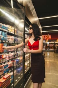 A stylish woman shops in a supermarket aisle, selecting yogurt from the freezer.