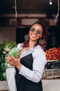 Fashionable woman smiling with sunglasses holding a vegetable at a bustling street market.