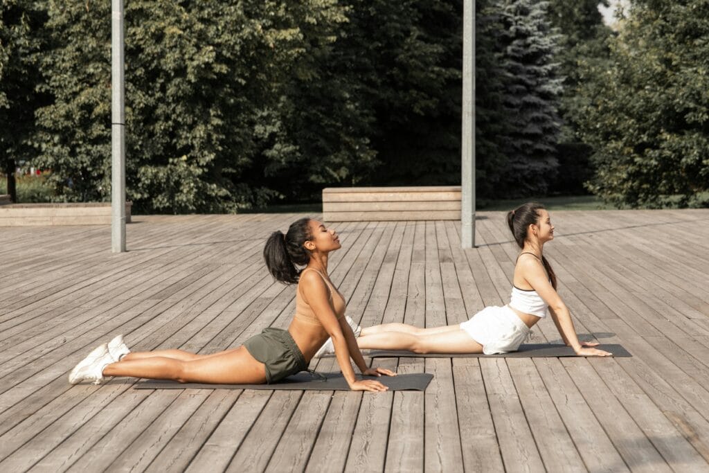 Two women practicing yoga outdoors in a tranquil park setting, focusing on flexibility and balance.
