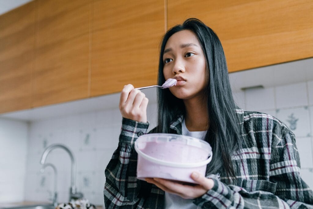 Asian woman enjoying ice cream in a casual indoor setting, looking pensive.