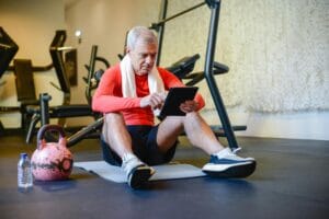 Senior man in a gym, seated on yoga mat, using a tablet for workout guidance.