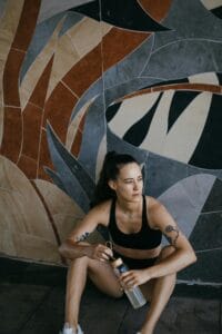 Young athletic woman in sports bra resting indoors with a water bottle, sitting against a mural wall.