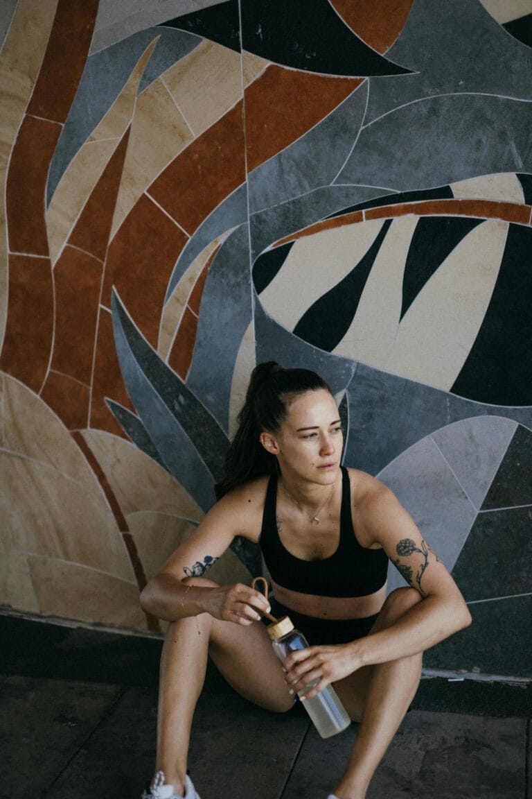 Young athletic woman in sports bra resting indoors with a water bottle, sitting against a mural wall.
