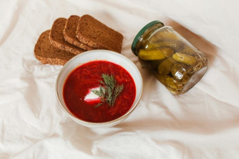 A hearty bowl of borscht with dill, accompanied by sliced brown bread and pickles on a white surface.