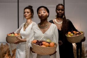 Three women in elegant attire holding baskets of fresh fruits, showcasing cultural elegance.