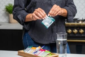 Close-up of hands holding a supplement package with pills next to a full glass of water in a kitchen setting.
