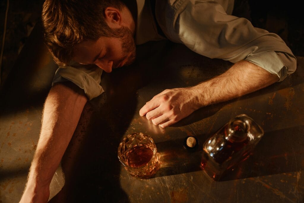 A man sleeps on a table with a whiskey glass and bottle nearby, suggesting exhaustion.