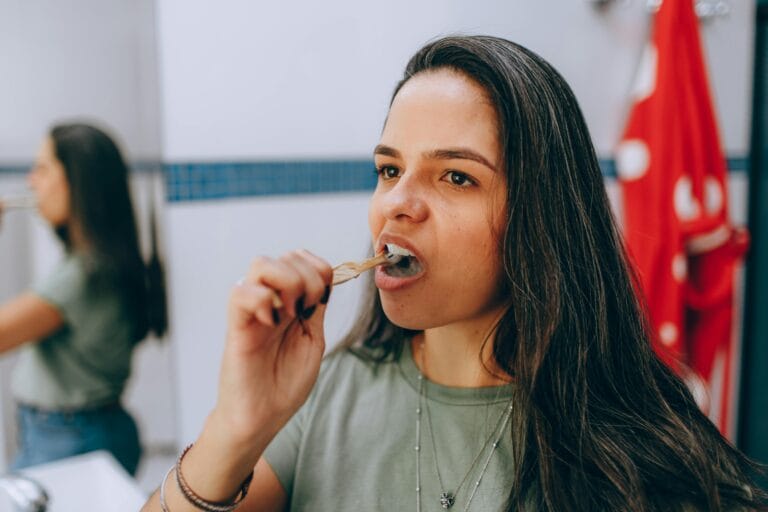 Woman brushing her teeth using a natural toothbrush in a bathroom, promoting oral hygiene and sustainability.