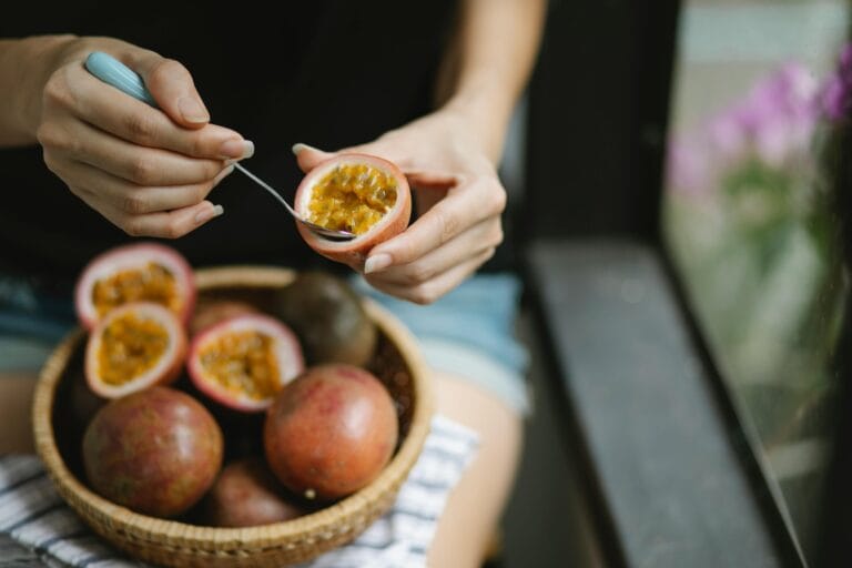 Crop lady in casual clothes sitting near window and eating fresh ripe passion fruits with small spoon and wicker plate on knees