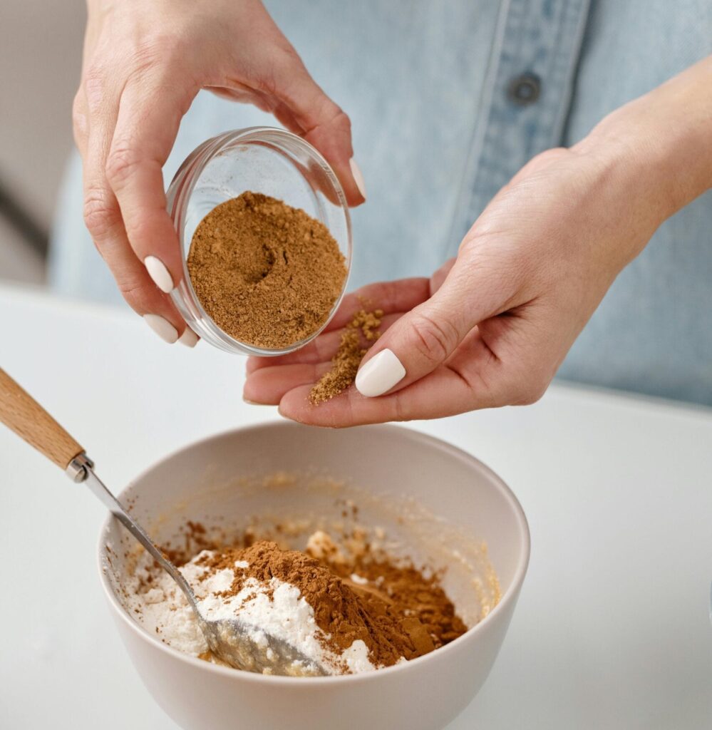 Close-up of hands adding cinnamon to a homemade baking mix in a modern kitchen setting.