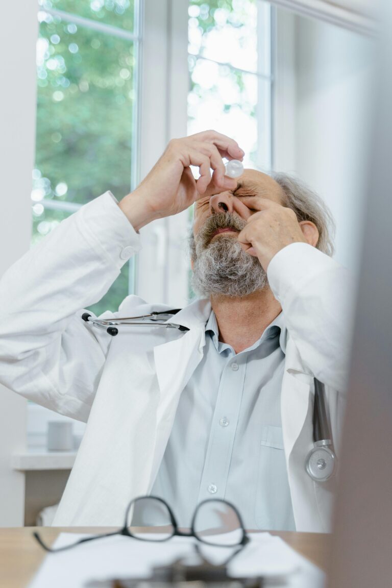 Senior male doctor in white coat applying eyedrops, sitting in a well-lit clinic.