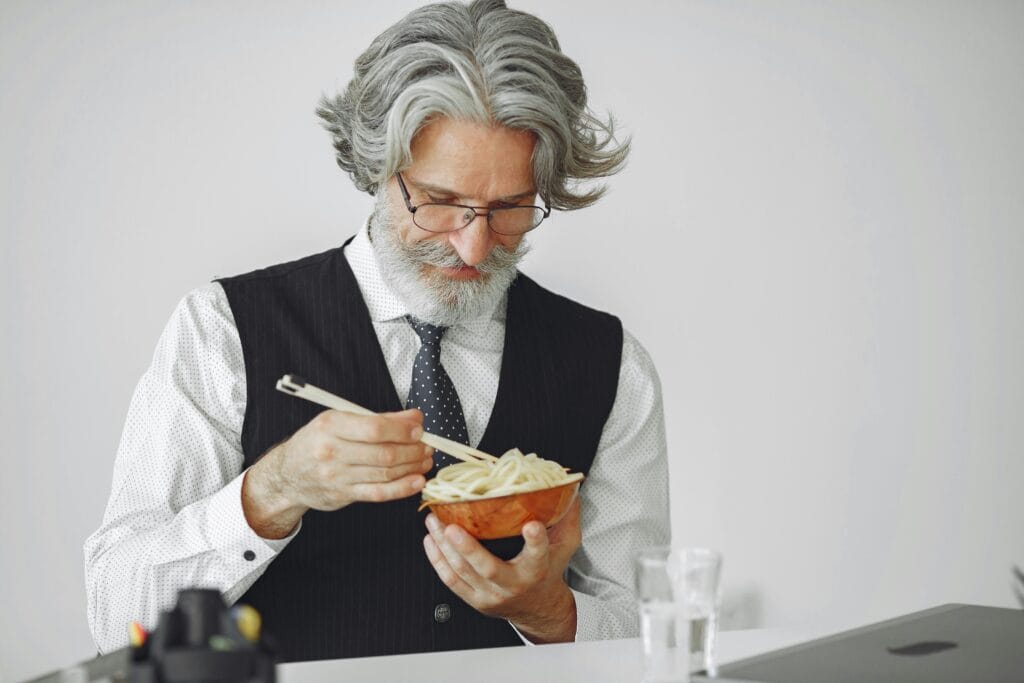 Elderly man with gray hair eating noodles with chopsticks, dressed in formalwear.