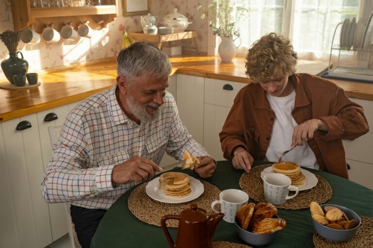 Elderly man and teenager sharing pancakes in a cozy kitchen setting, enjoying quality time together.