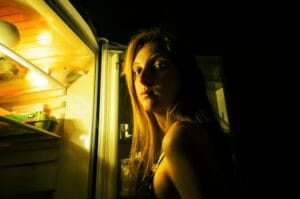 A woman looking thoughtfully into an open fridge at night, illuminated by its light.