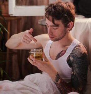 Tattooed man sitting in bedroom holding a jar of honey, creating a relaxed indoor atmosphere.